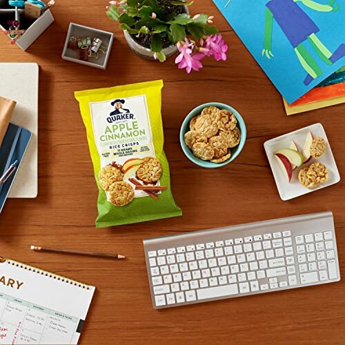 Desk with Quaker apple cinnamon rice crisps, keyboard, plant, calendar, and snacks.